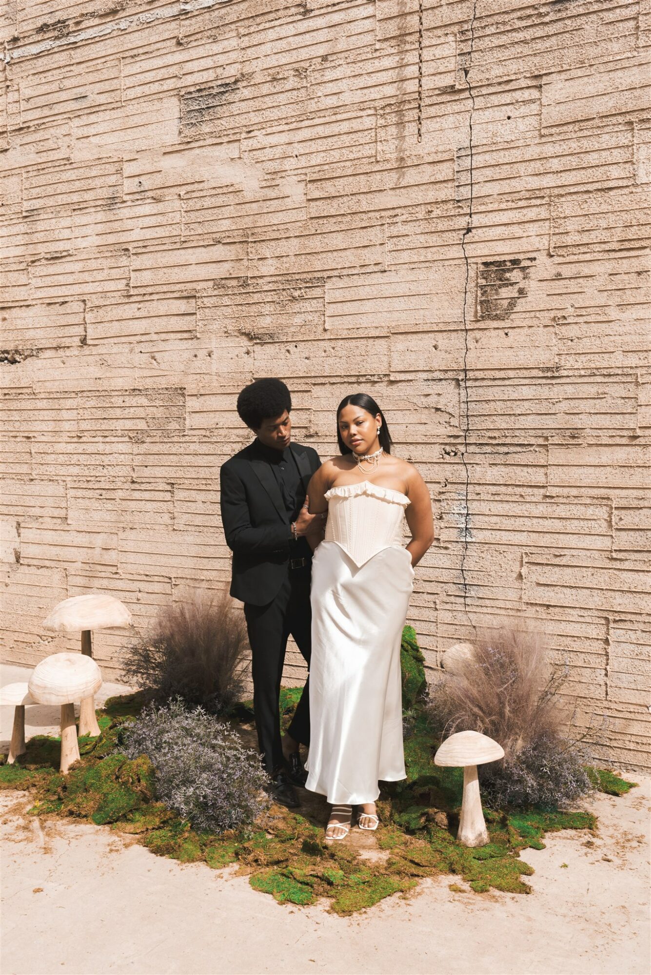A man and woman in formal attire standing outdoors against a textured brick wall, surrounded by plants and mushrooms.