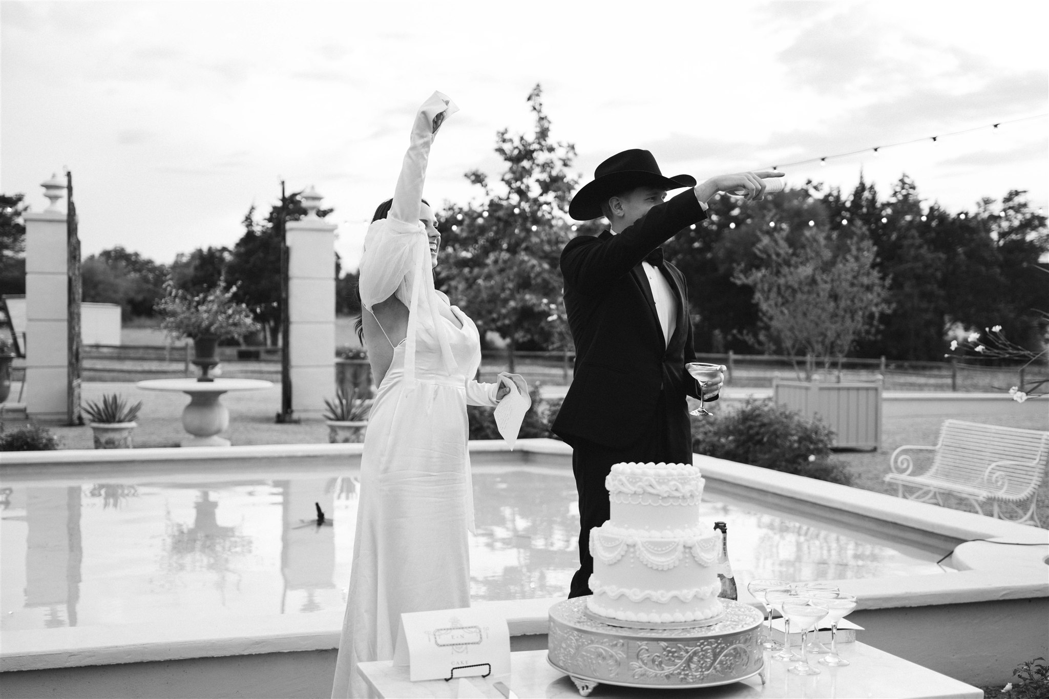 Two women in dresses and hats stand near a cake on a table outdoors, with a pool and trees in background.