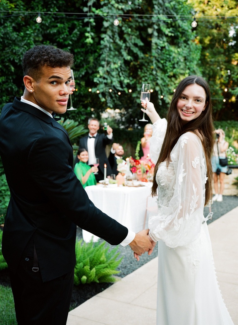 Young couple holding hands at outdoor celebration, smiling, with string lights and guests in background.