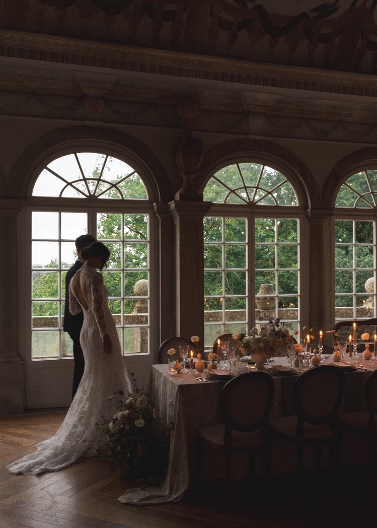 Bride in wedding dress standing near large arched windows with a decorated table nearby.
