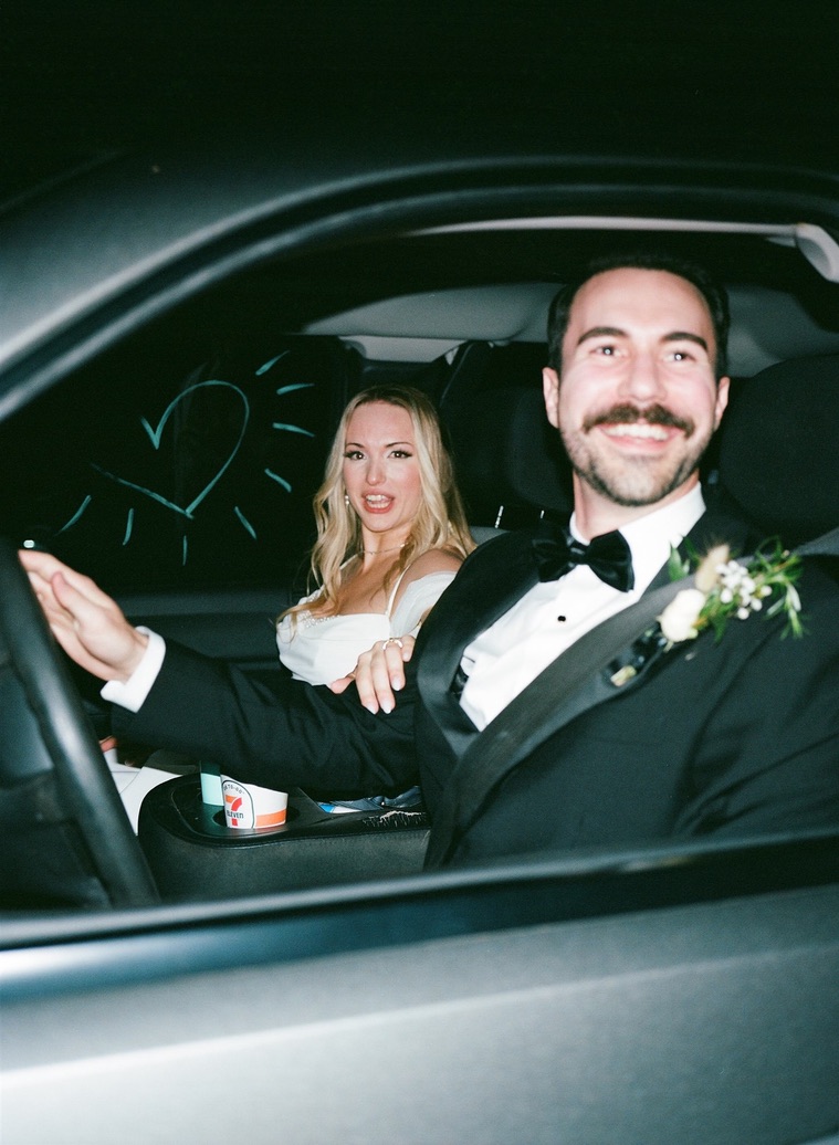 Man and woman in a car, man in tuxedo smiling, woman with blonde hair, heart drawing on window.