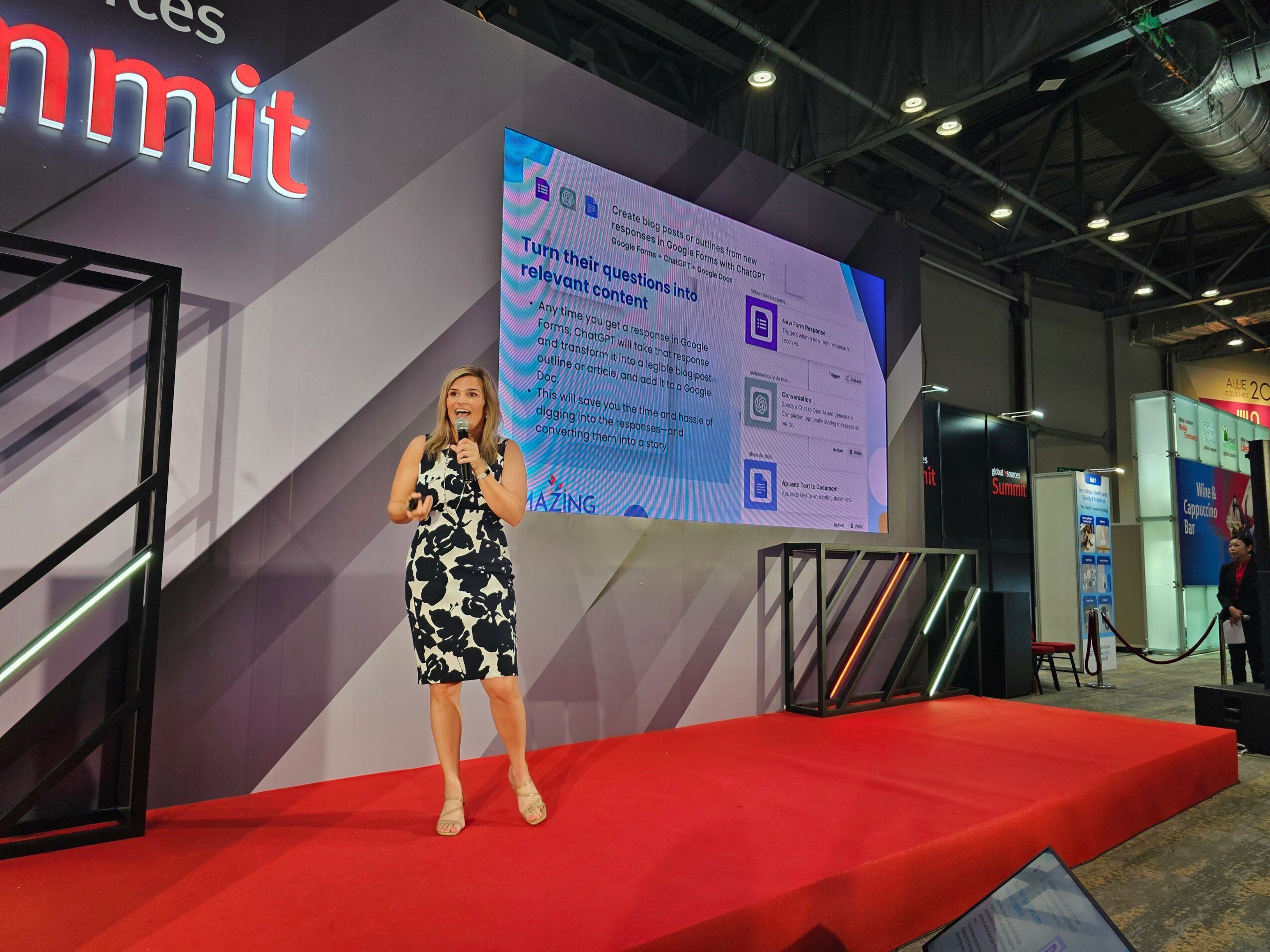 Woman in a black and white floral dress speaking on stage with a large screen behind her at a conference.
