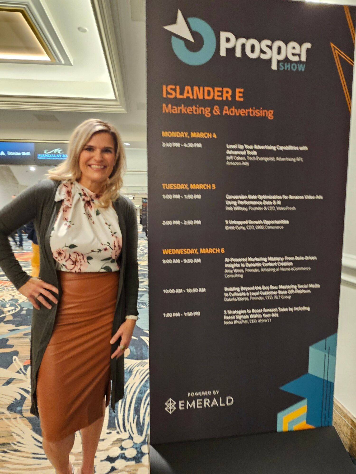 Woman with blonde hair in floral blouse and brown skirt standing next to a large event poster.