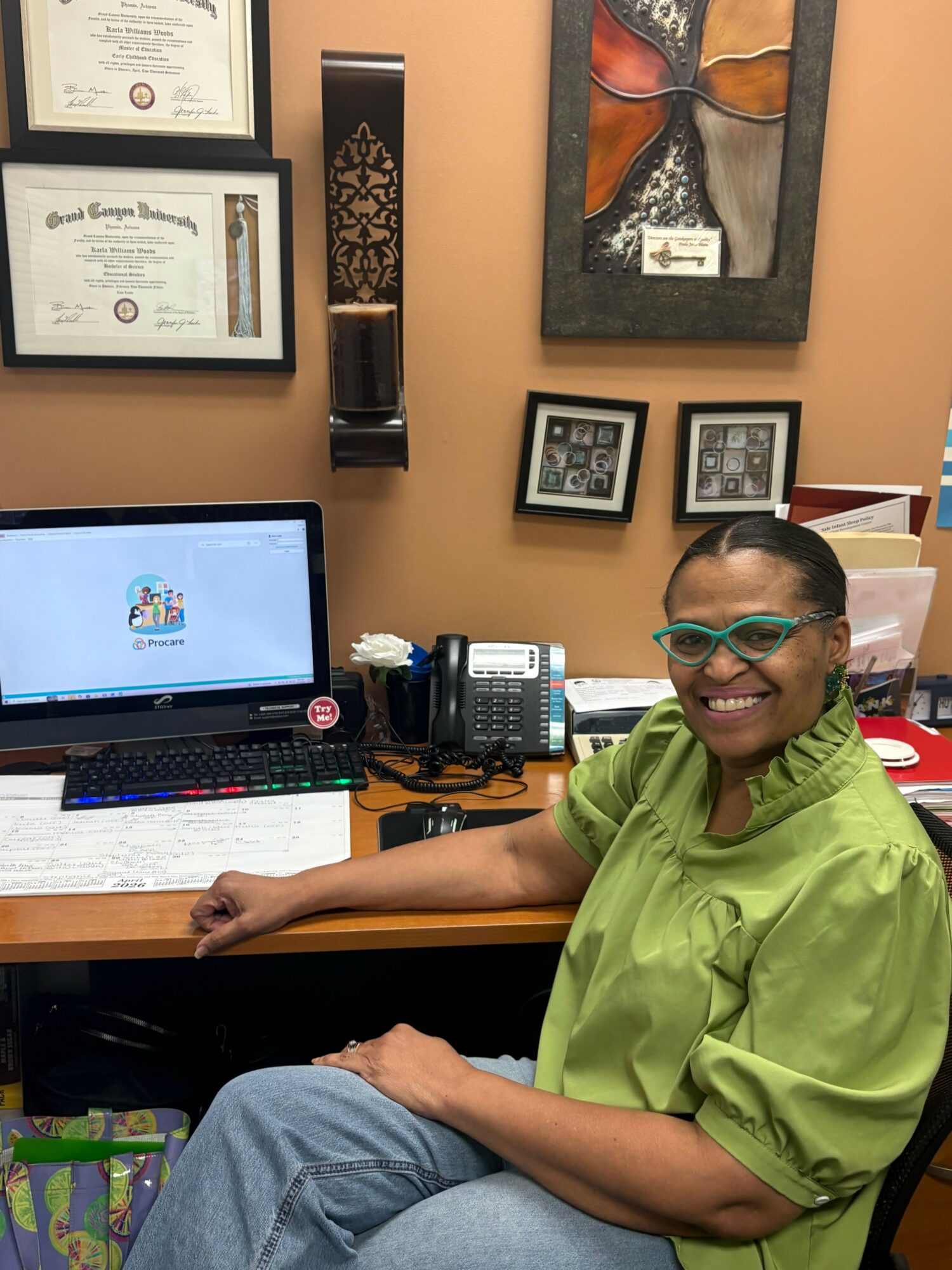 Smiling woman with glasses in green shirt sitting at desk with computer, certificates, and framed artwork on wall behind her.