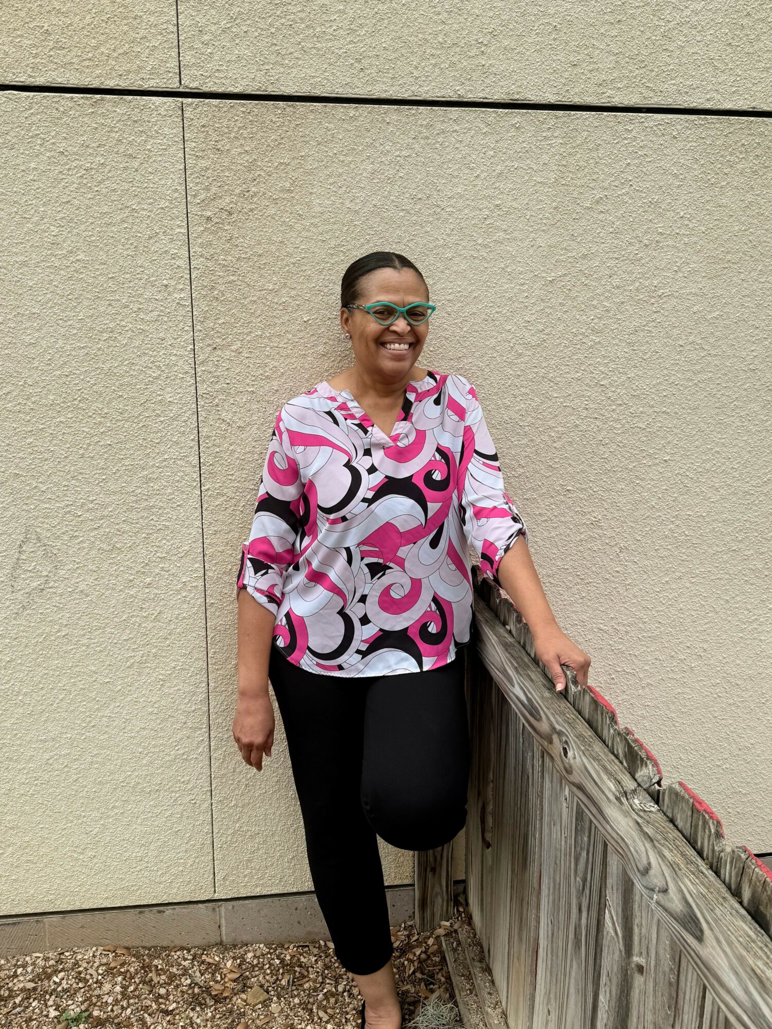 Smiling woman with glasses in a pink, white, and black patterned blouse, black pants, standing outdoors near a wooden fence.