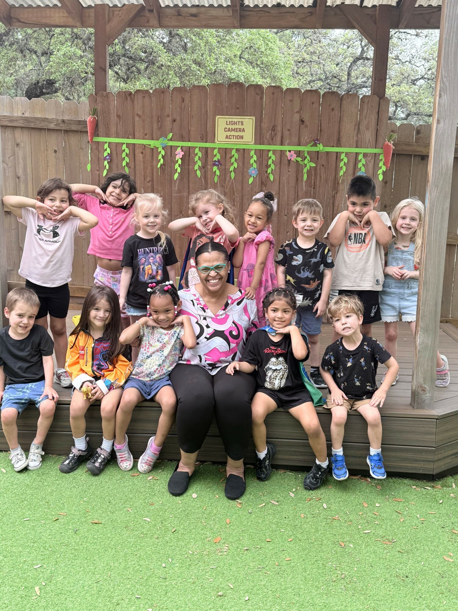 Group of children and adult sitting and standing outdoors on a wooden platform, smiling and posing for the photo.