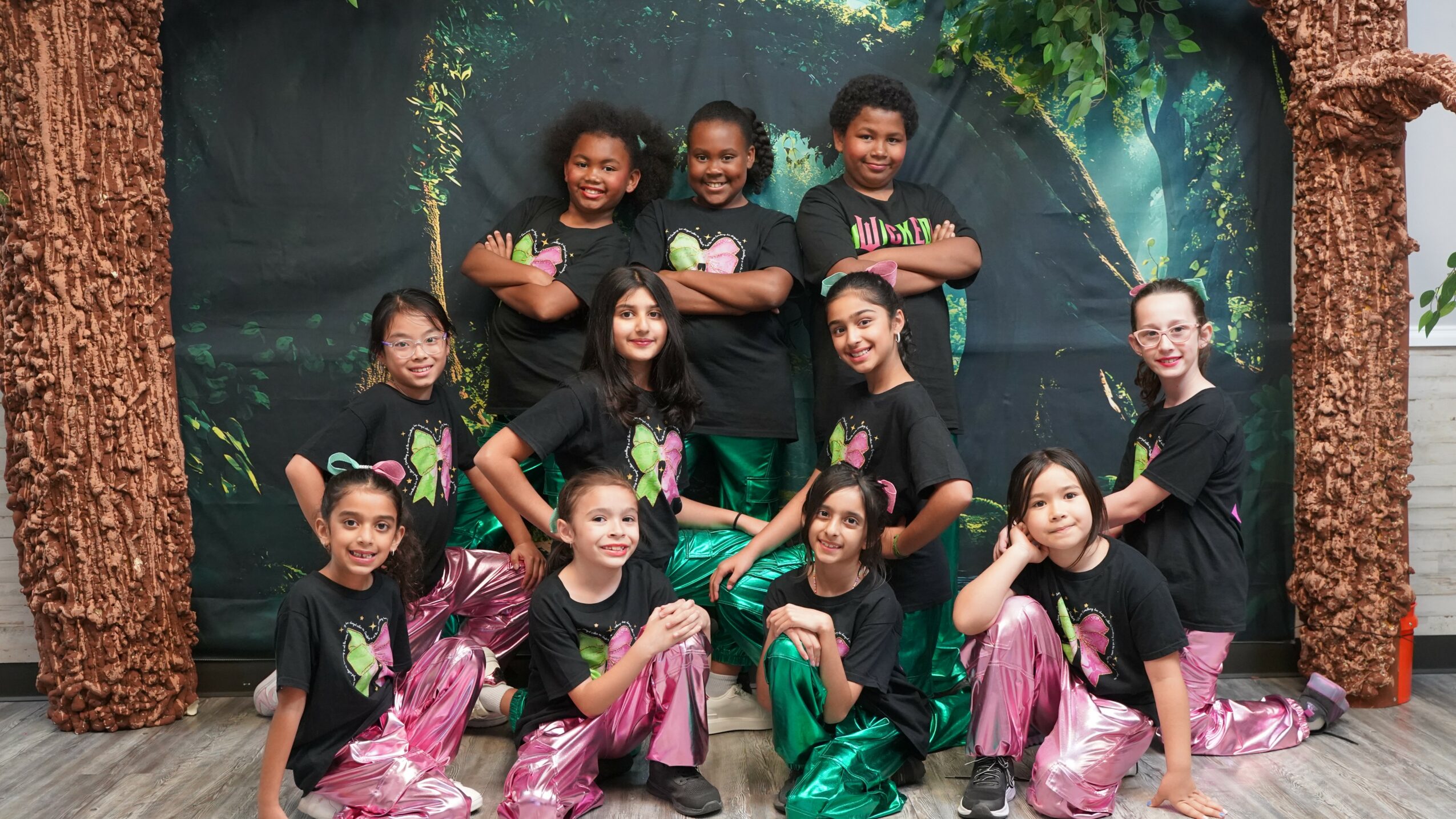 Group of ten children in black shirts and colorful pants, posing in front of a forest backdrop.