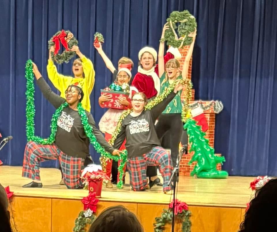 Group of people on stage celebrating with Christmas decorations and costumes, some holding presents, in front of a blue curtain.