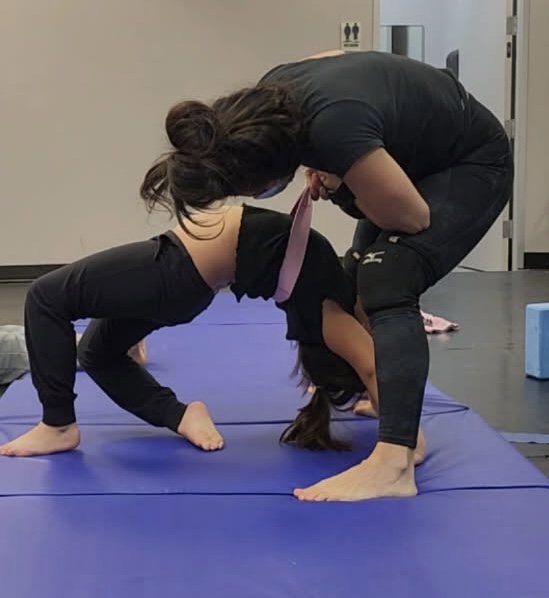 Two women practicing yoga on a blue mat, one bending backward and the other assisting by holding her waist.