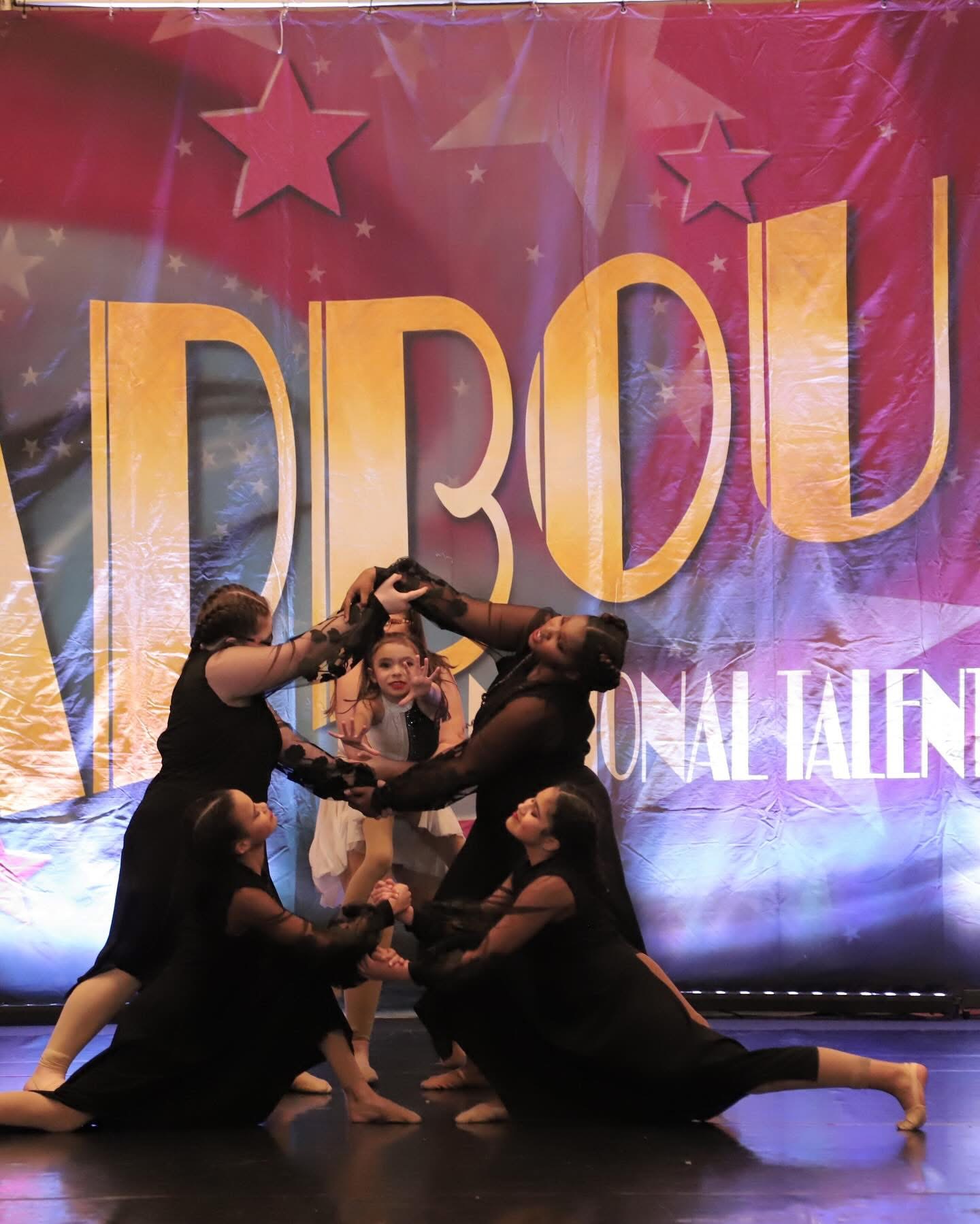 Group of five young girls performing a dance on stage with a colorful backdrop and large text.