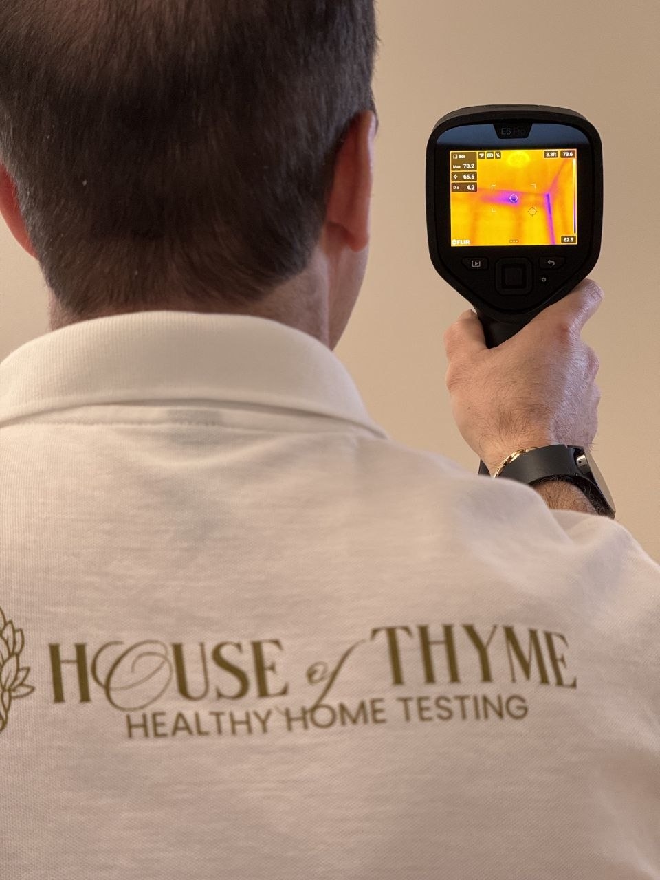 Person wearing a white shirt with 'HOUSE of THYME' and 'HEALTHY HOME TESTING' text, holding a thermal imaging camera.