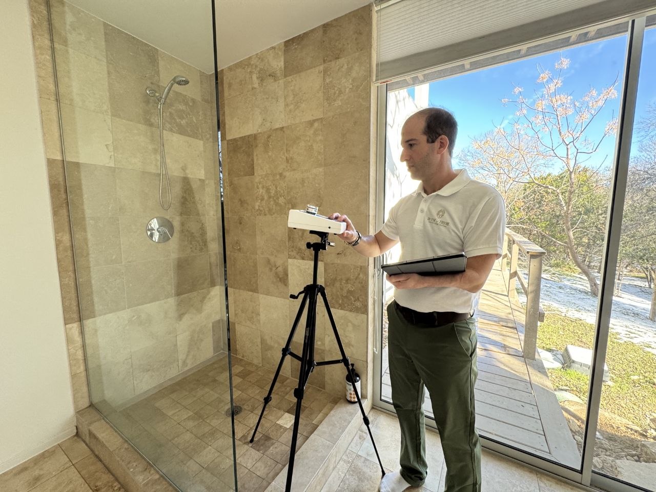 Man standing inside a room with a large window, holding a clipboard and a device, with a camera on a tripod.