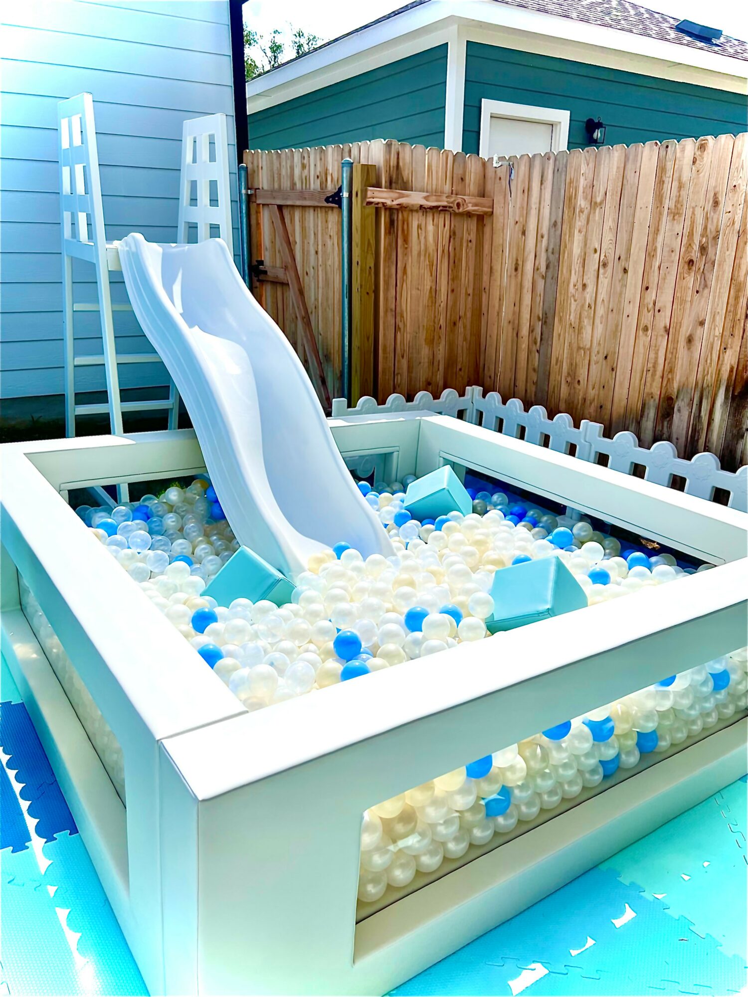 Small outdoor ball pit with a slide, filled with white, blue, and clear balls, in a backyard setting.
