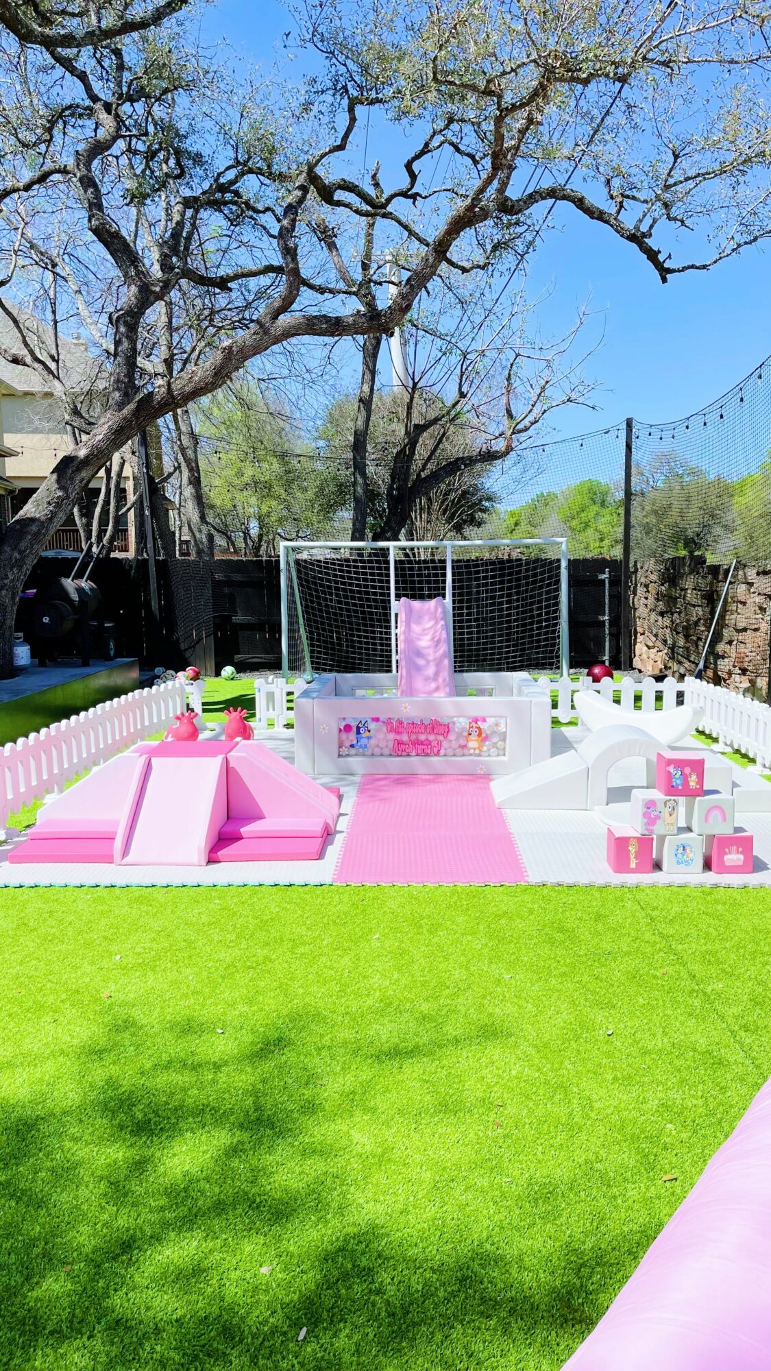 Outdoor party setup with pink and white decorations, seating, and a soccer goal in the background under a large tree.