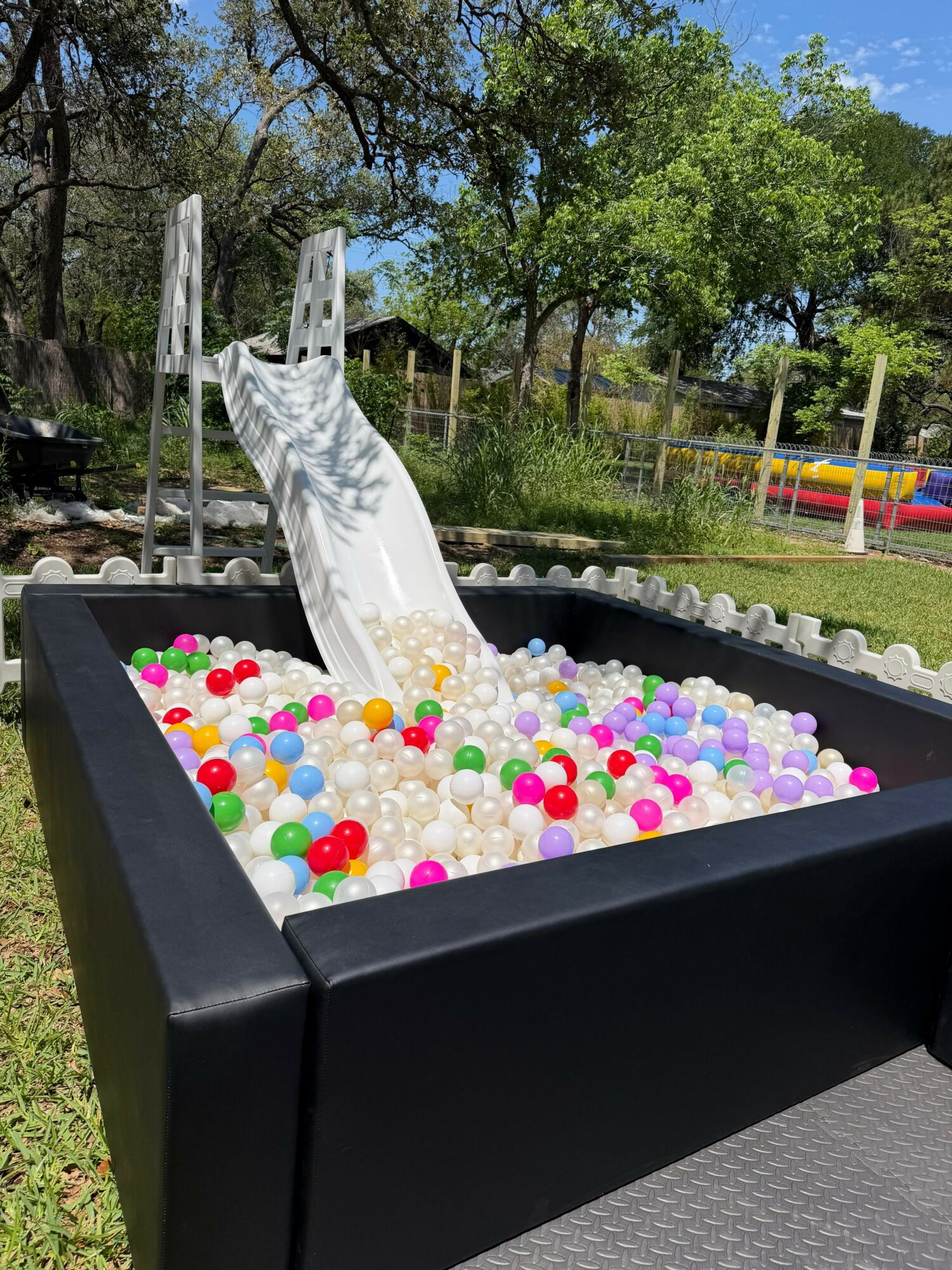 Ball pit with colorful plastic balls and a slide in an outdoor setting with trees and a fence.