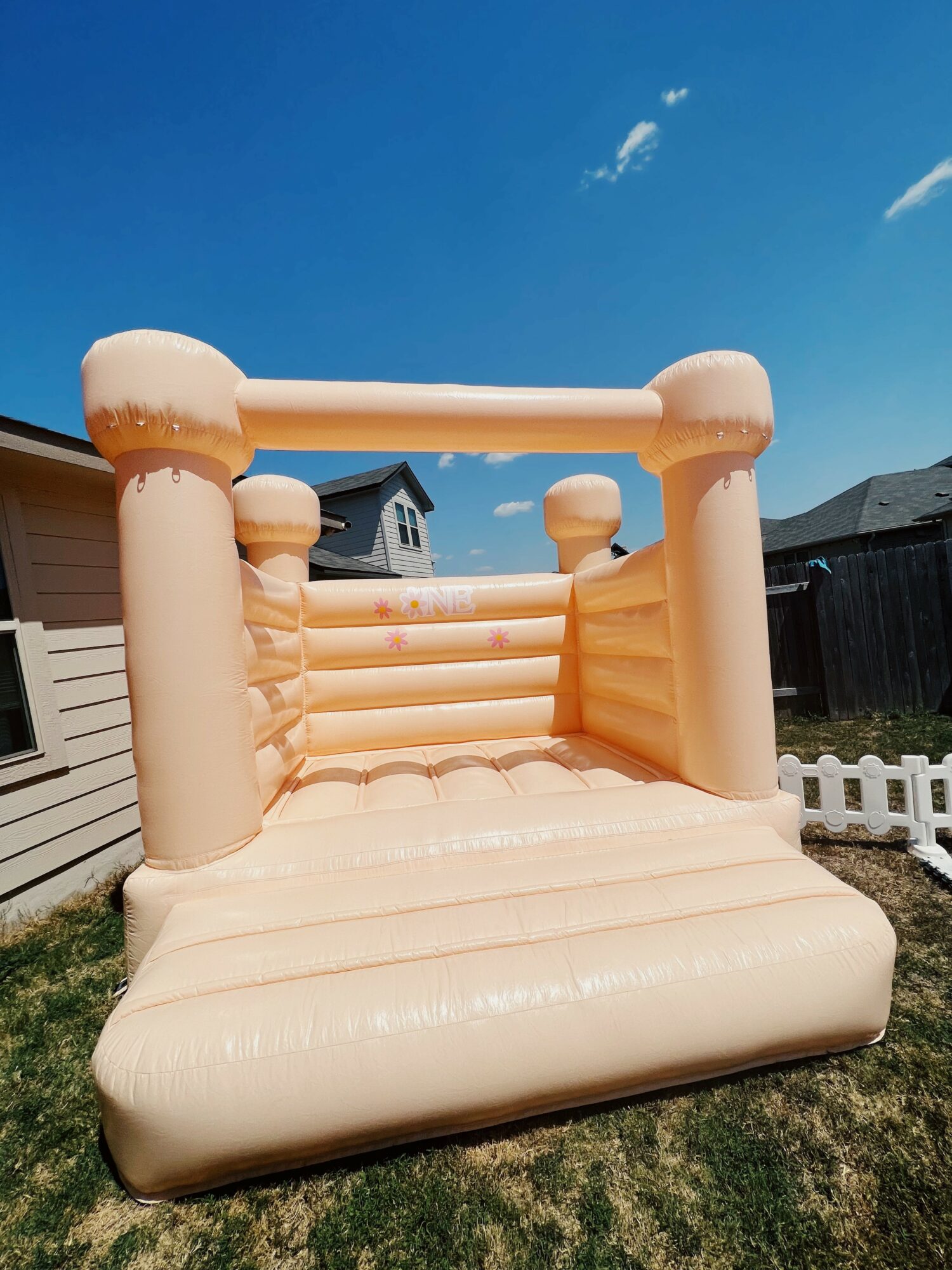Inflatable bounce house with beige color, four tall pillars, and a small entrance, set on grass under a blue sky.