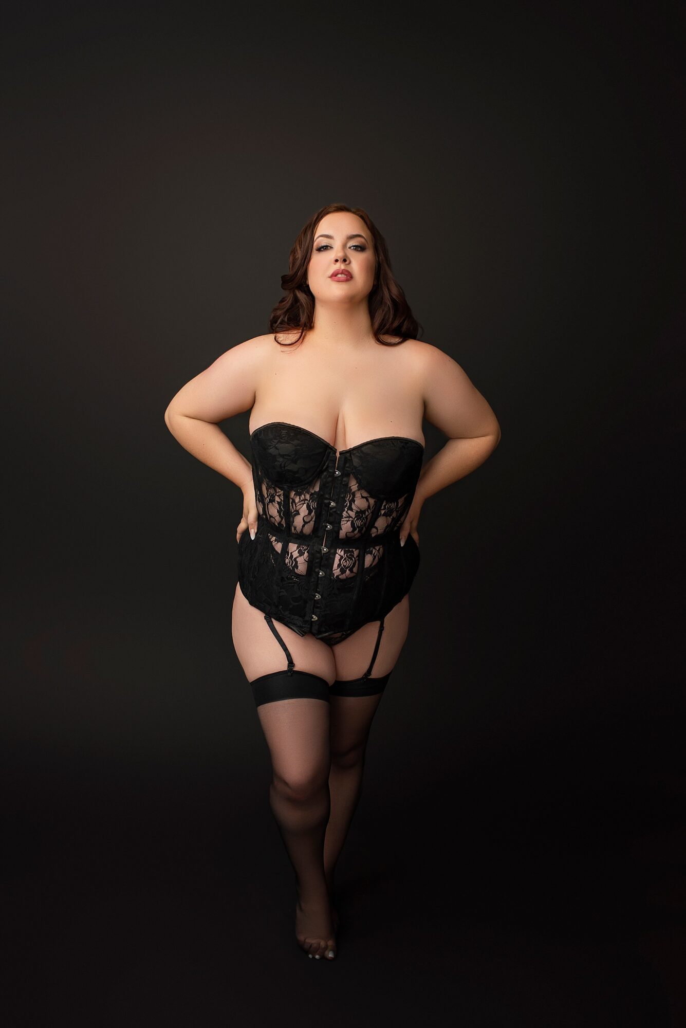 Woman in black lace corset and stockings standing against black background.