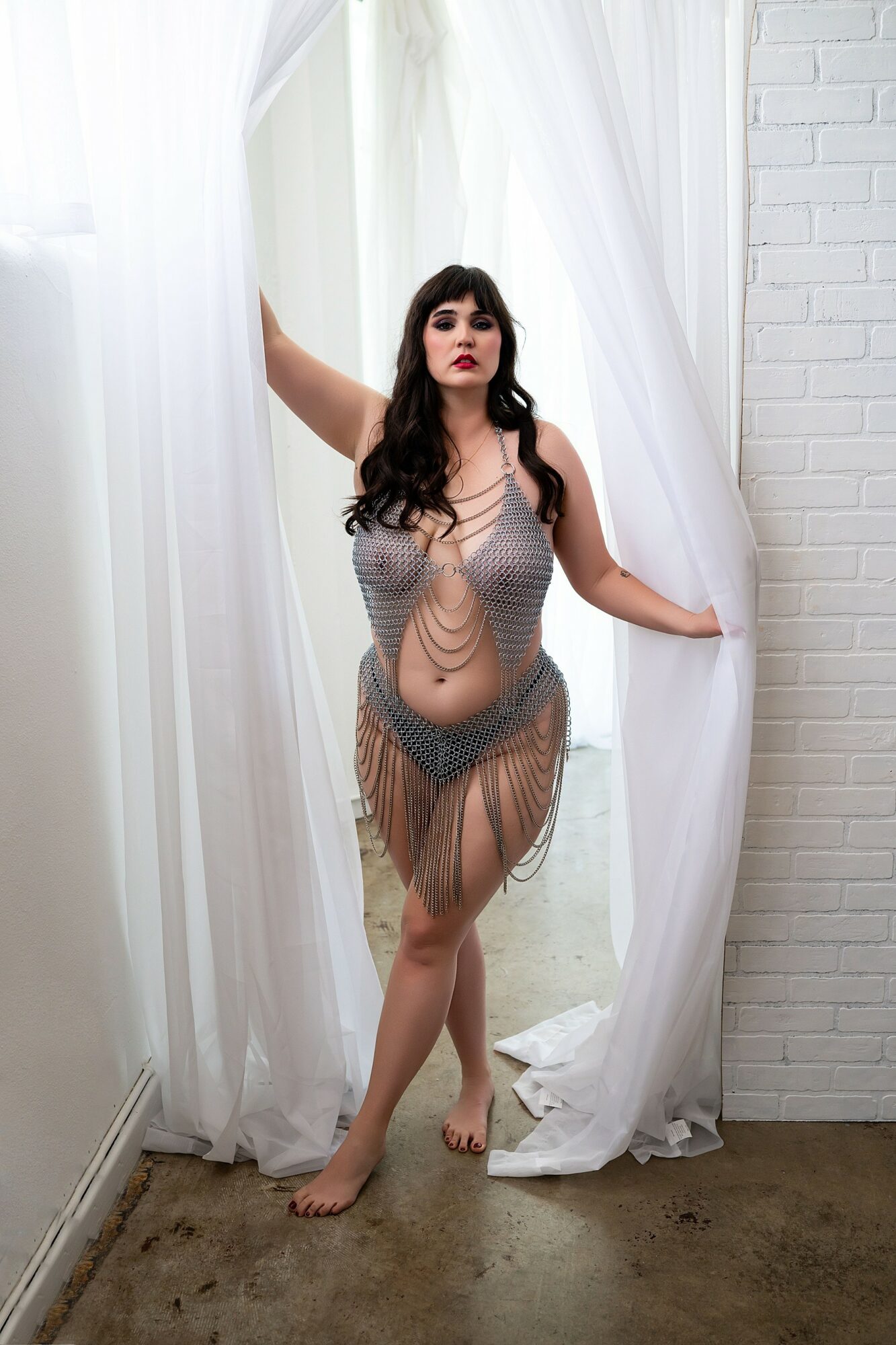 Woman with long dark hair and red lipstick stands in doorway with white curtains, wearing a sheer, fringed dress.