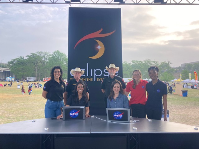 Six people standing behind a table with NASA laptops, outdoors with a large banner and tents in the background.