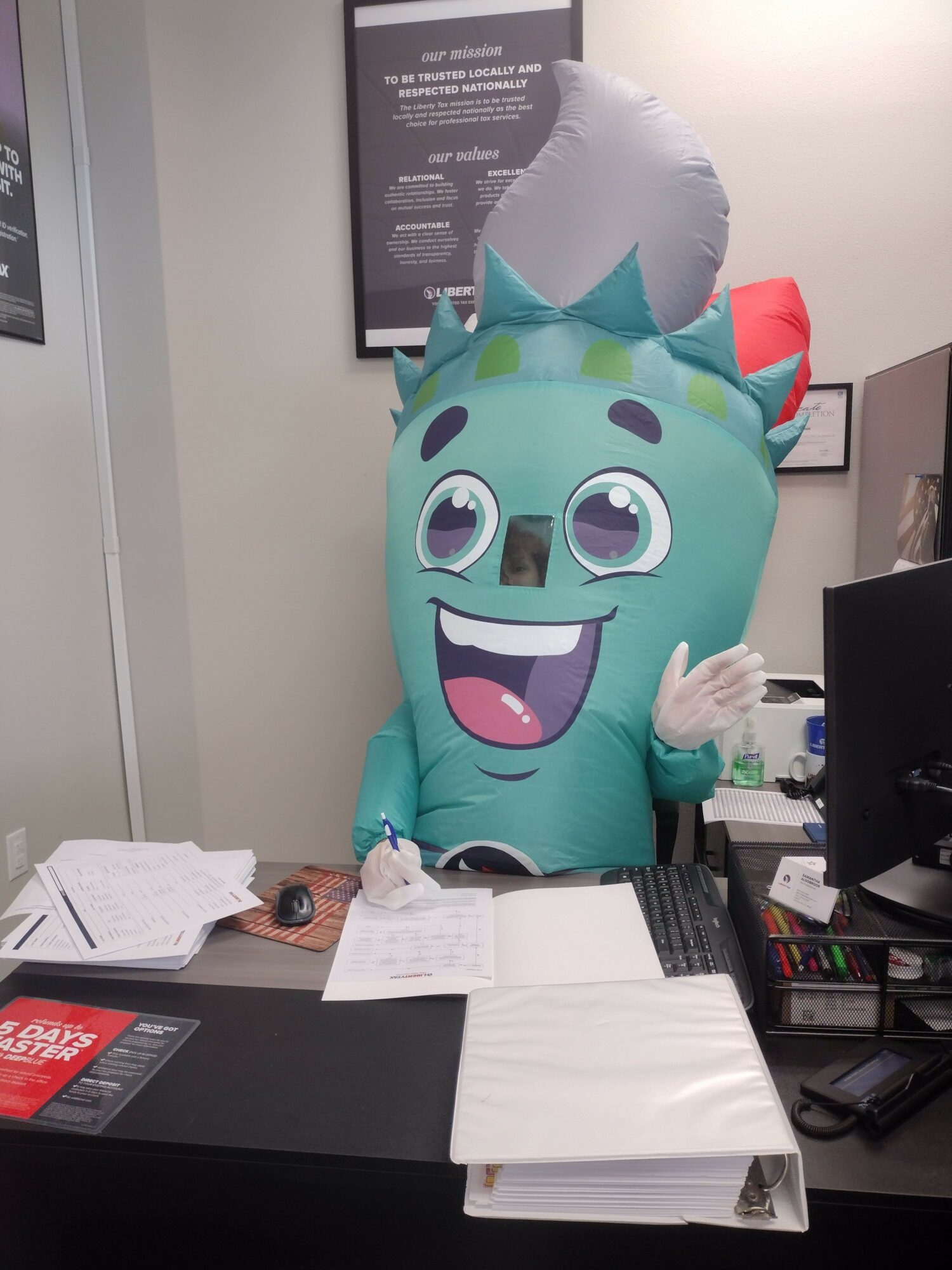 Person in a cartoonish monster costume with large eyes and a big smile, waving hand, in office setting.