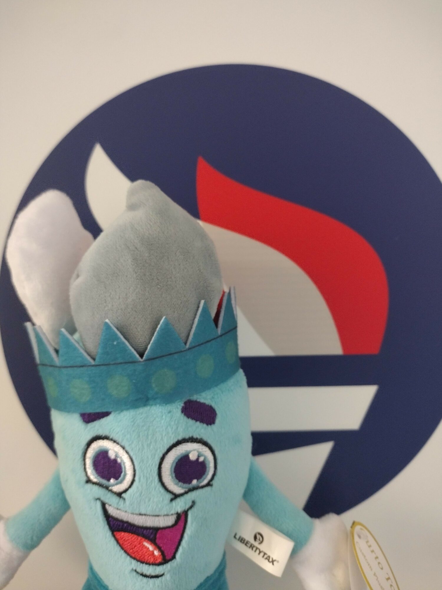 Plush toy with a crown and large eyes, smiling, in front of a circular logo with red, white, and blue elements.