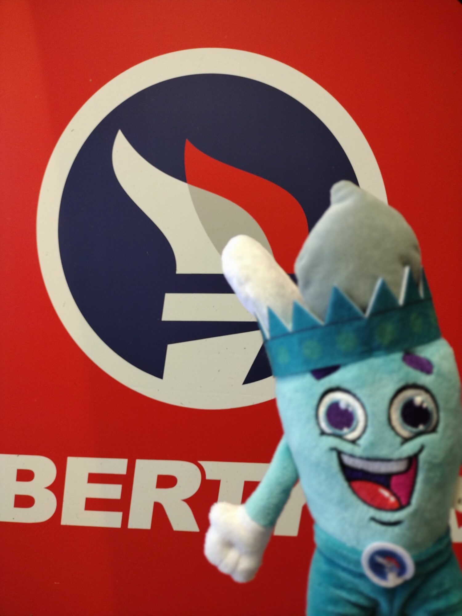 A plush toy character with a crown and big eyes in front of a logo with a flame and a circle.