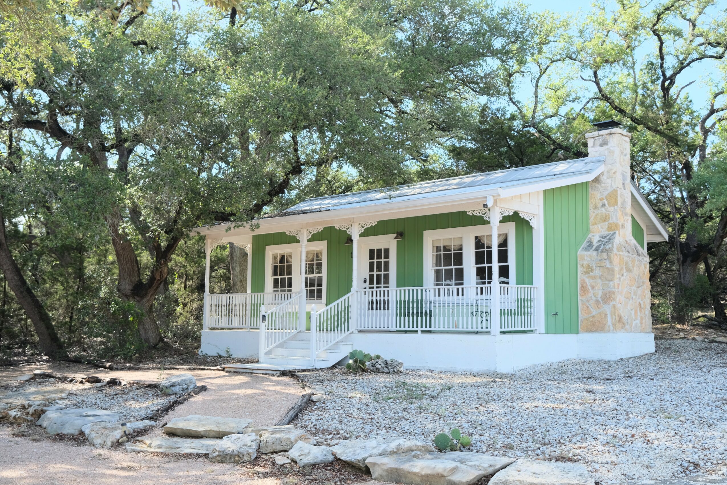 Small house with porch, green walls, white railing, surrounded by trees and gravel ground.