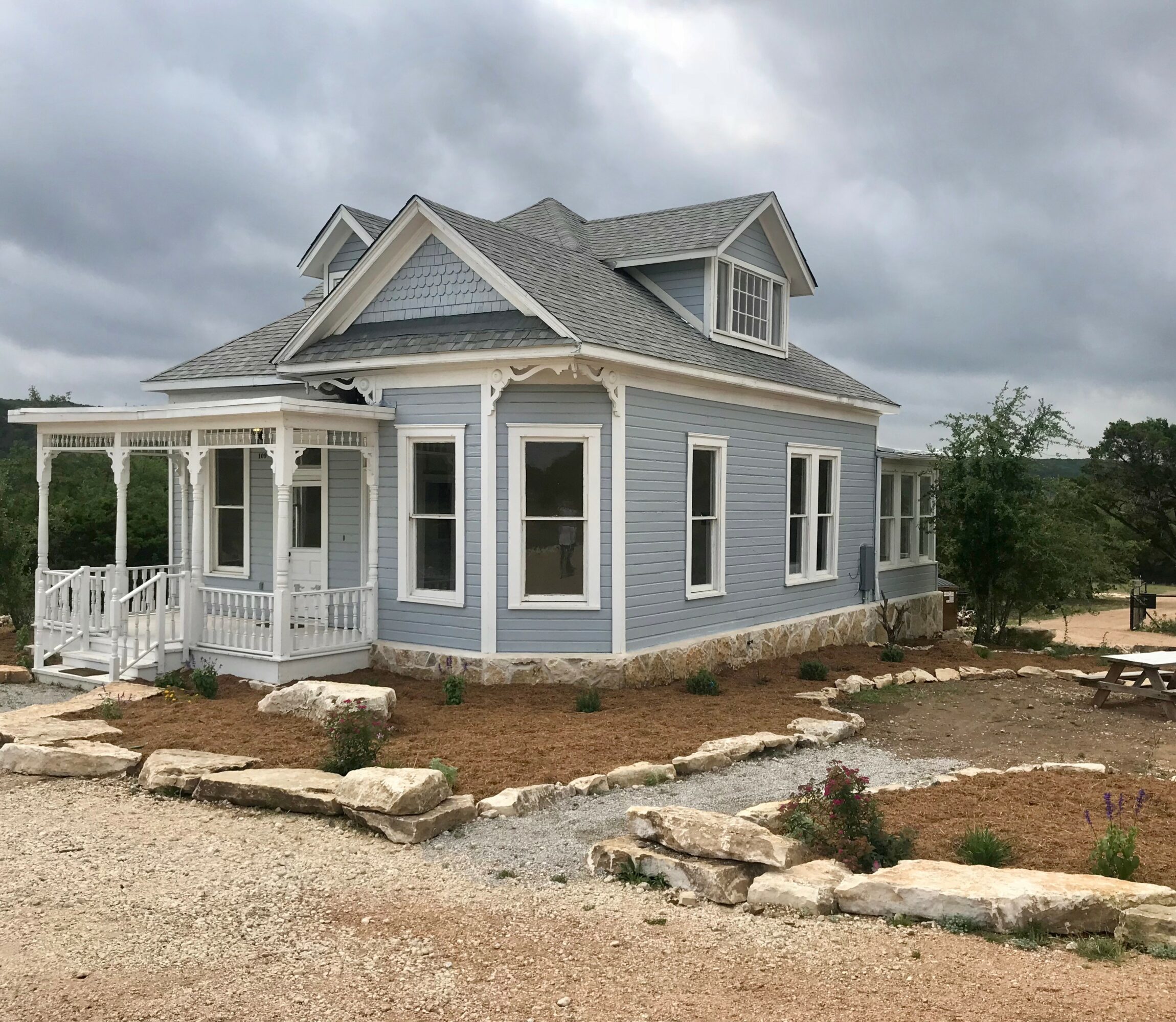 Blue house with white trim, porch, multiple windows, and a gabled roof, under cloudy sky, surrounded by rocks and dirt.