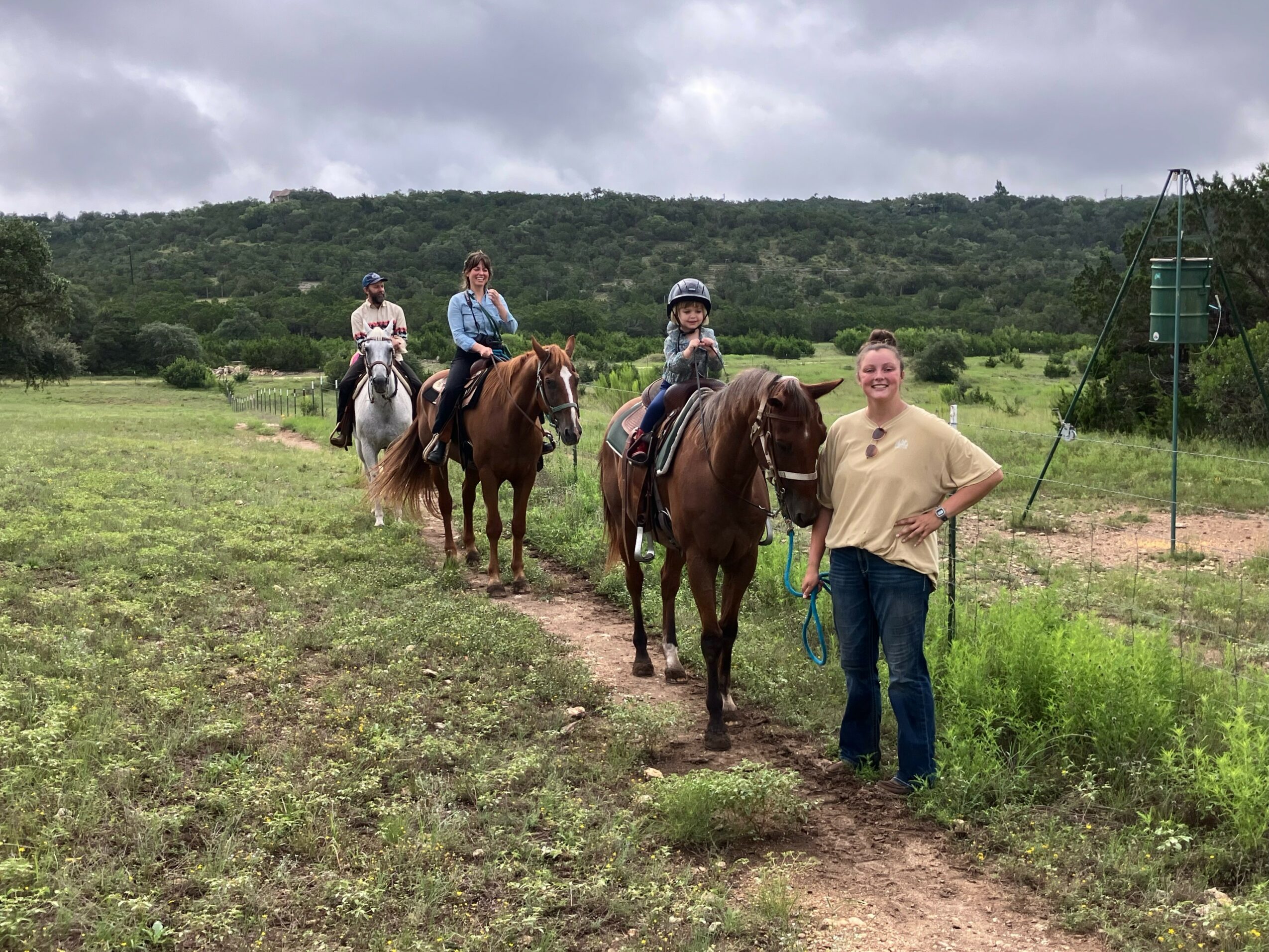 Four people on horseback and one person standing on a trail in a grassy field with hills and cloudy sky.