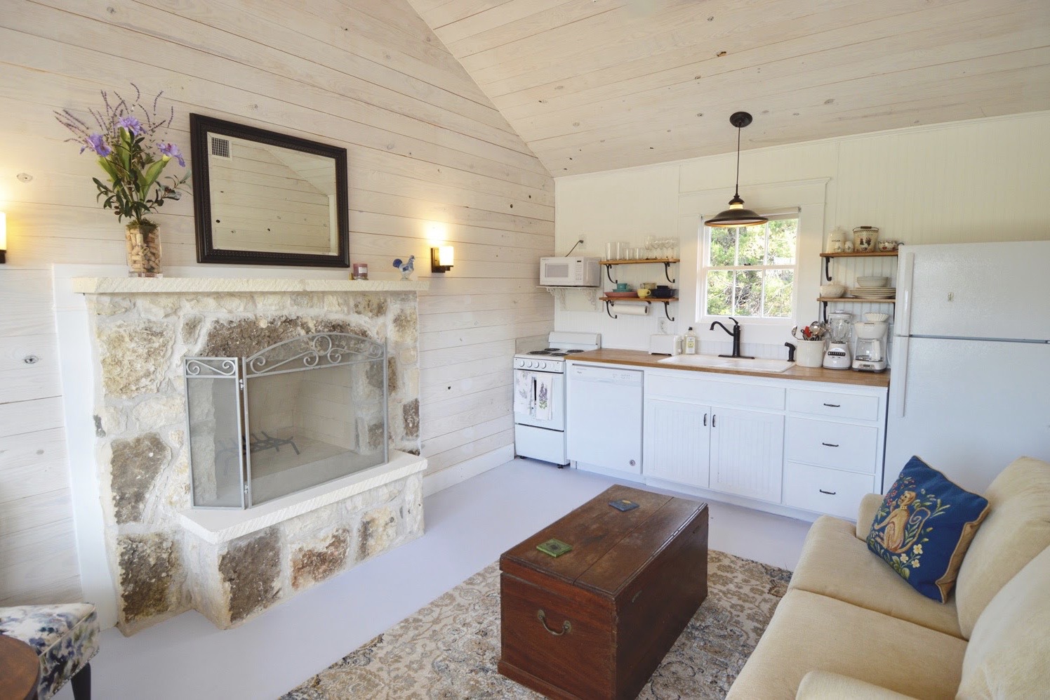 Living room with fireplace, white cabinets, refrigerator, window, and sofa with pillows, wooden chest, and rug.