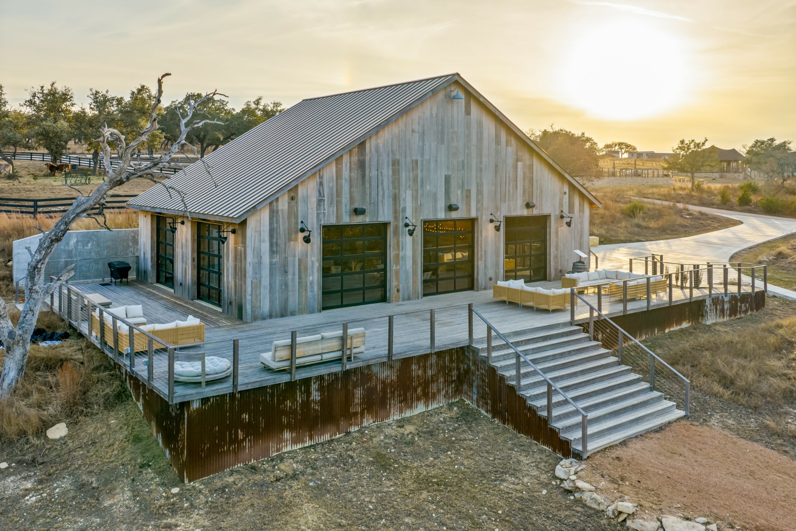Wooden barn with three garage doors on a dock over water, stairs leading up, sunset in background.