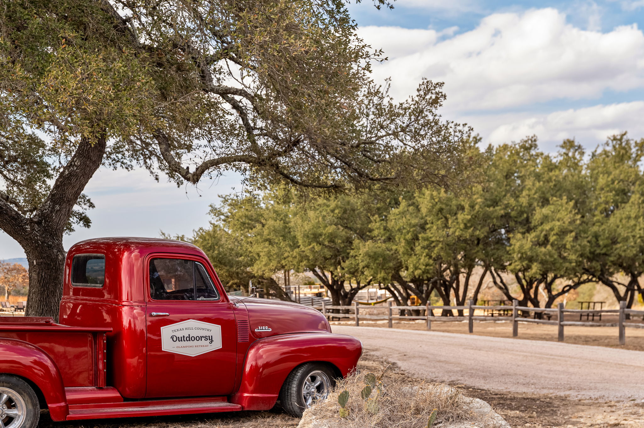 Red vintage pickup truck parked near a tree with green leaves, fence, and a dirt road under a partly cloudy sky.