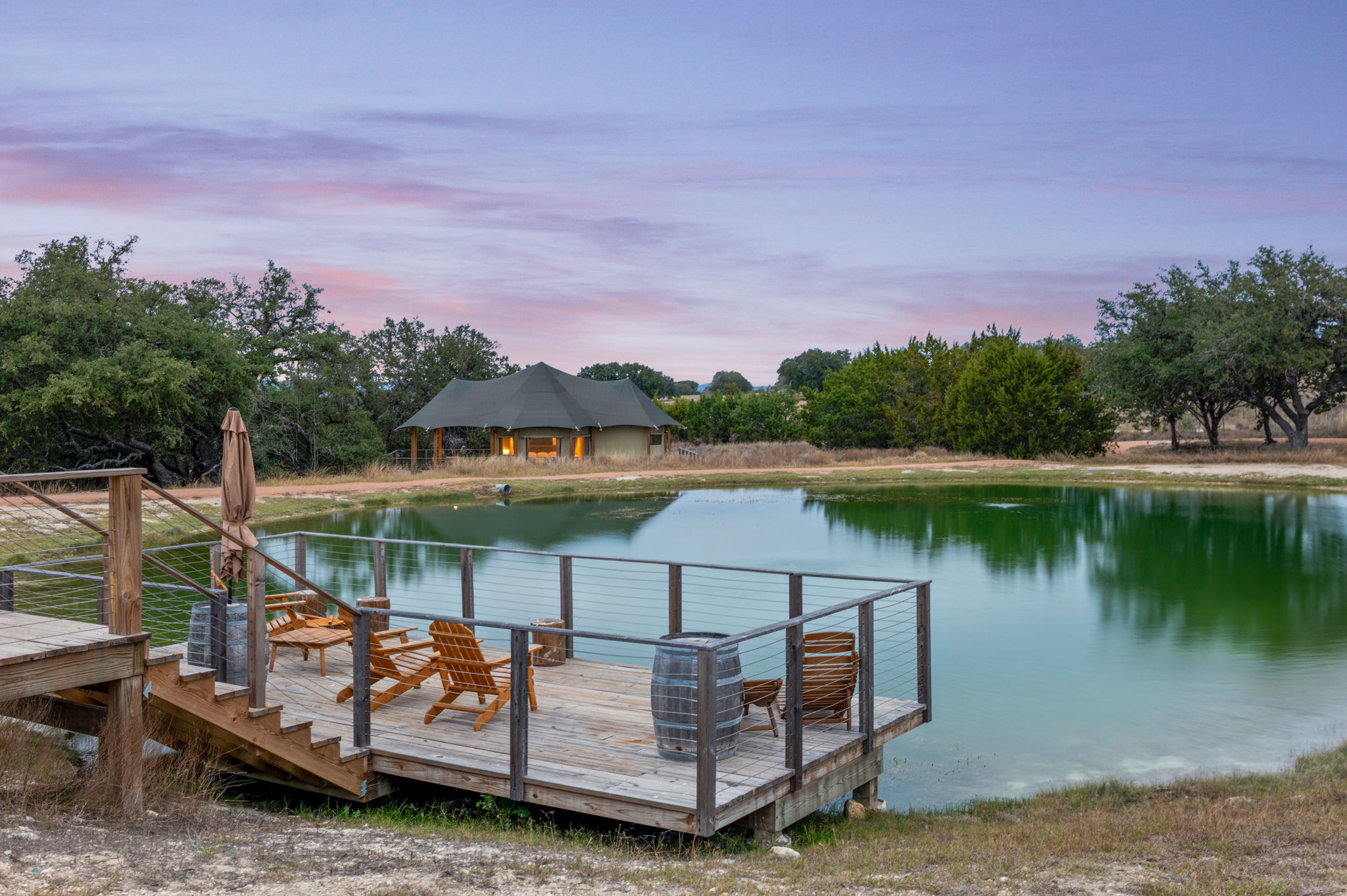 Wooden deck with chairs and umbrella overlooking a calm lake with trees and a building in the background.