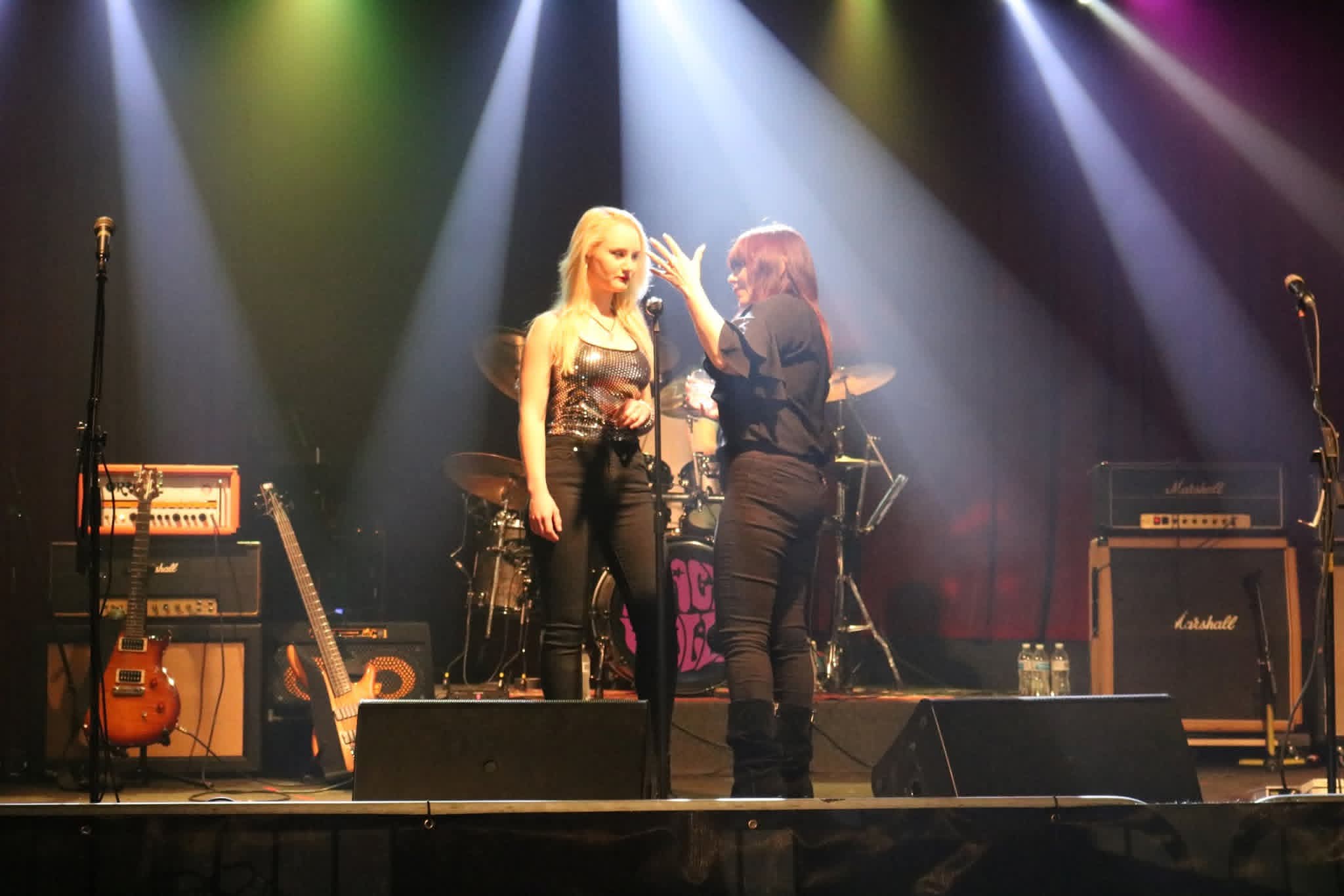 Two women on stage with musical instruments and equipment, illuminated by colorful stage lights.