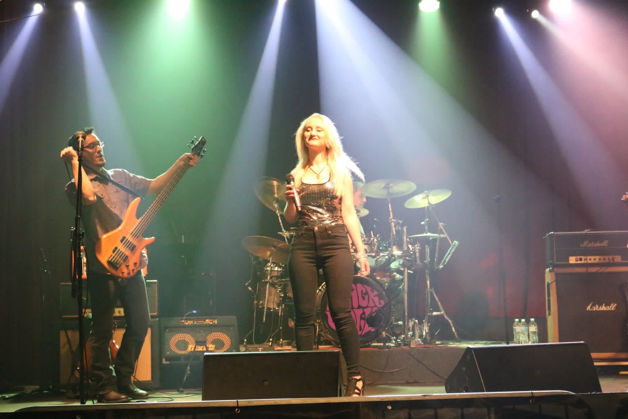 Two musicians perform on stage with colorful lighting, one singing and the other playing guitar, with drums and amplifiers in background.