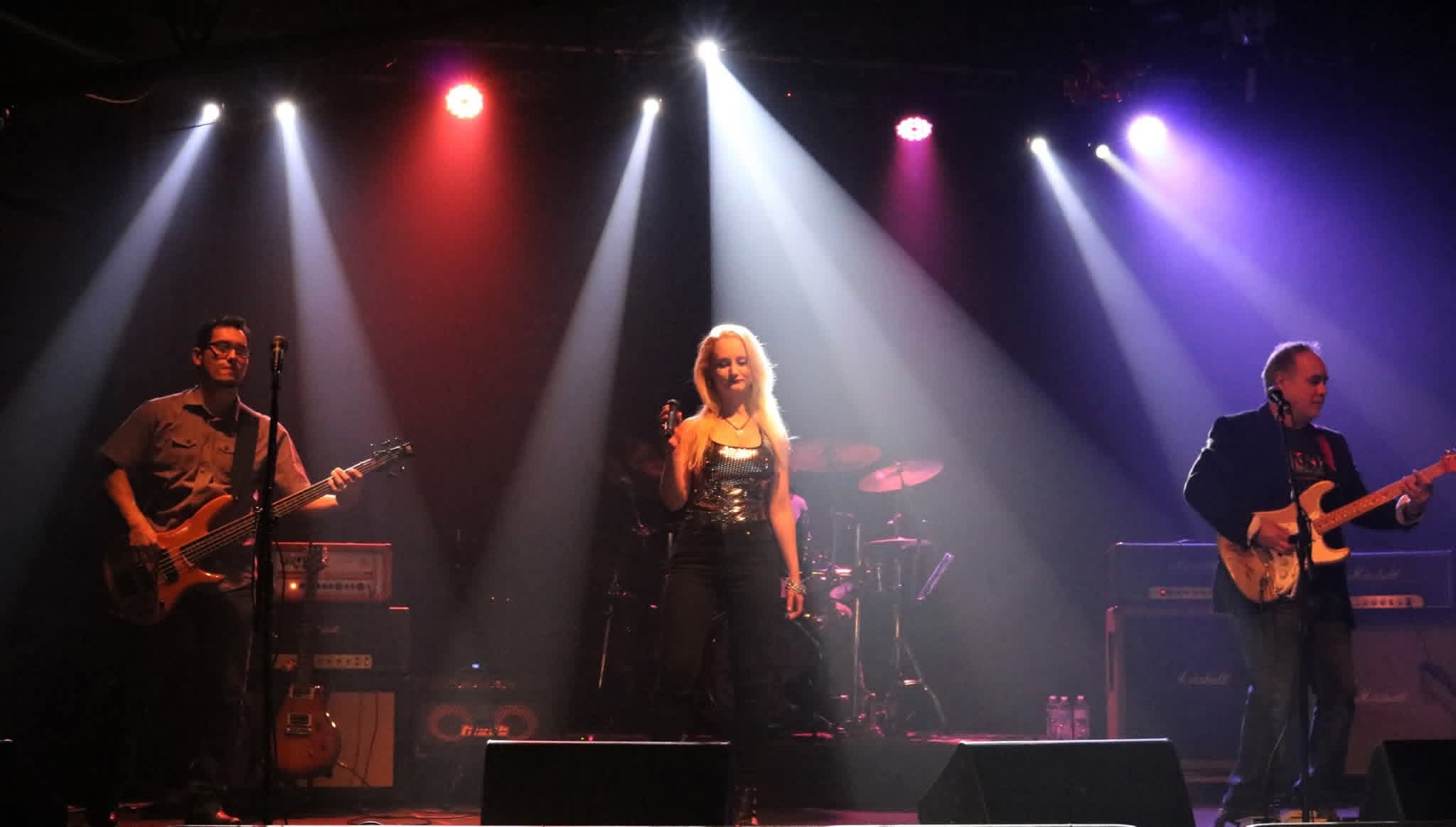 Three musicians perform on stage with colorful spotlights overhead, a female singer in the center, two guitarists on sides.