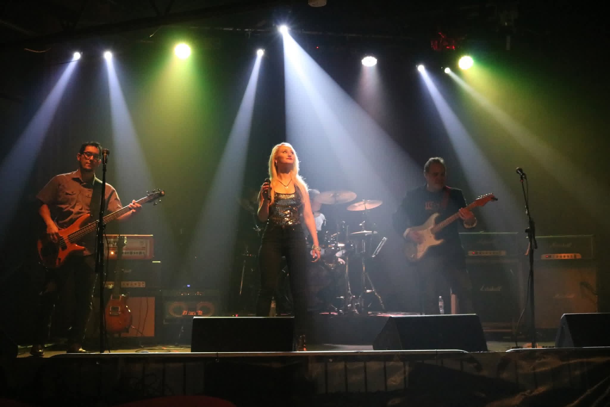 Four musicians perform on stage with colorful spotlights shining down, one singer in the center, two guitarists on sides, drummer behind.