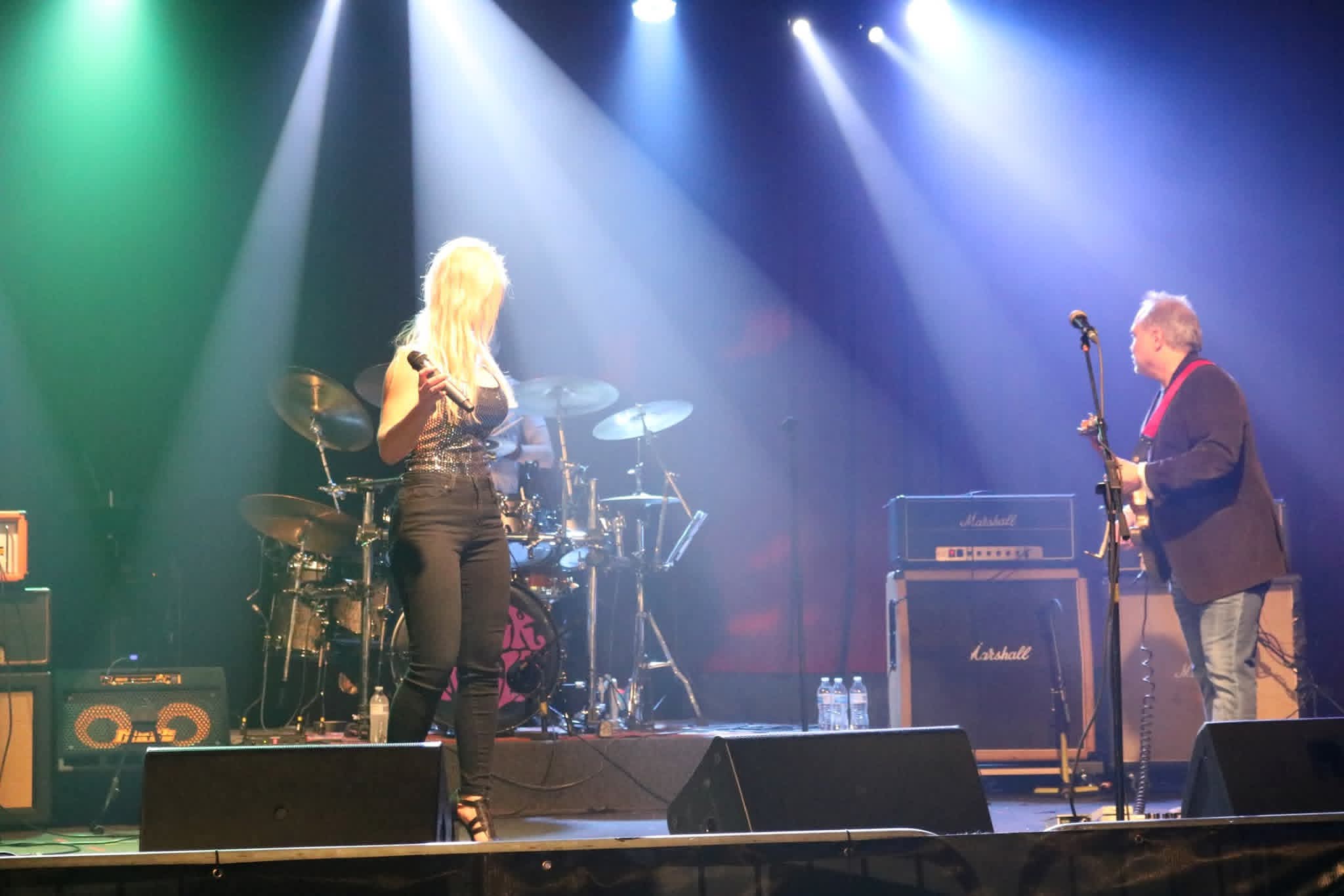 Two musicians perform on stage with colorful lighting, one singing and the other playing guitar, with drums in the background.