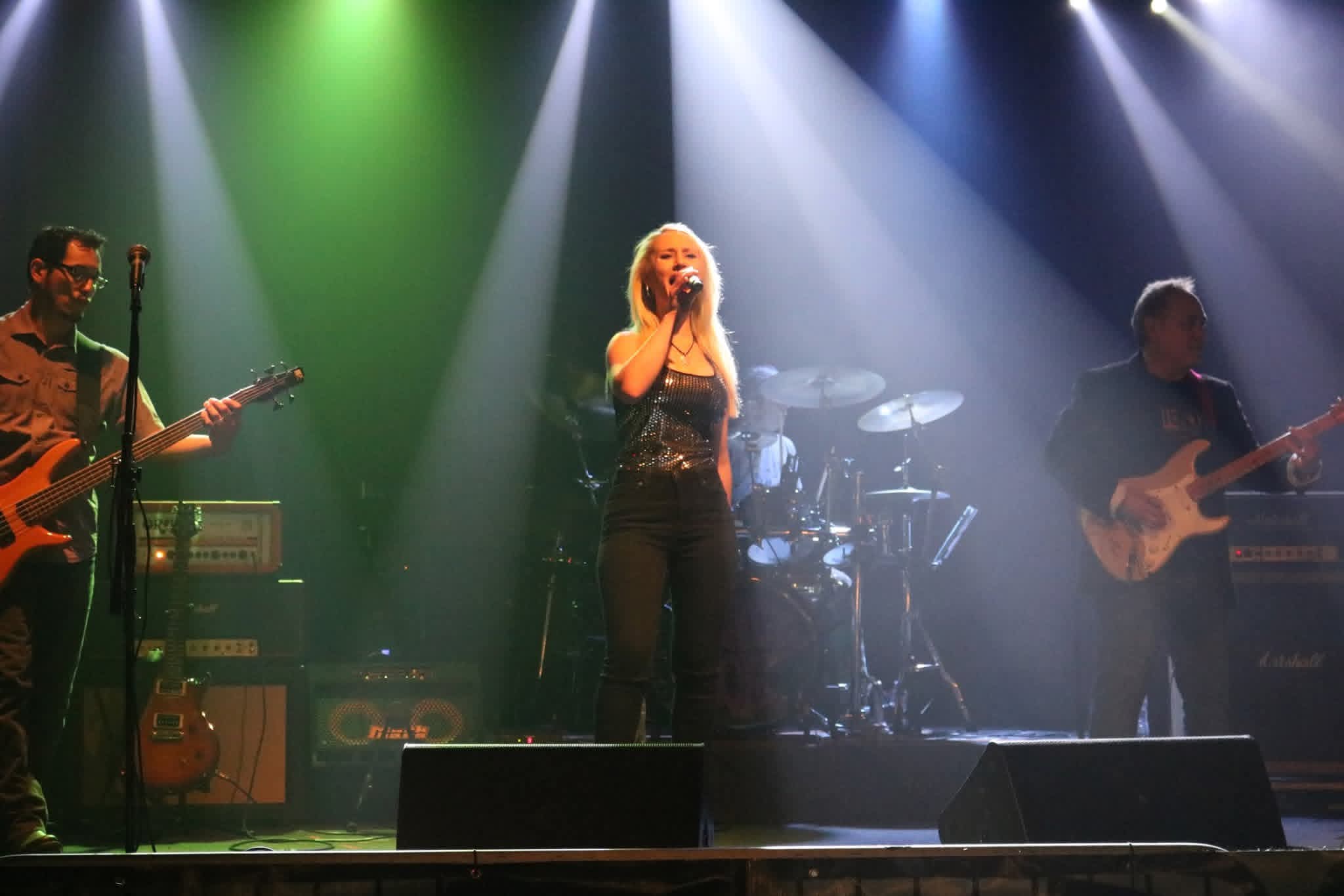 Four musicians perform on stage with colorful lighting, including a singer in the center, two guitarists, and a drummer.