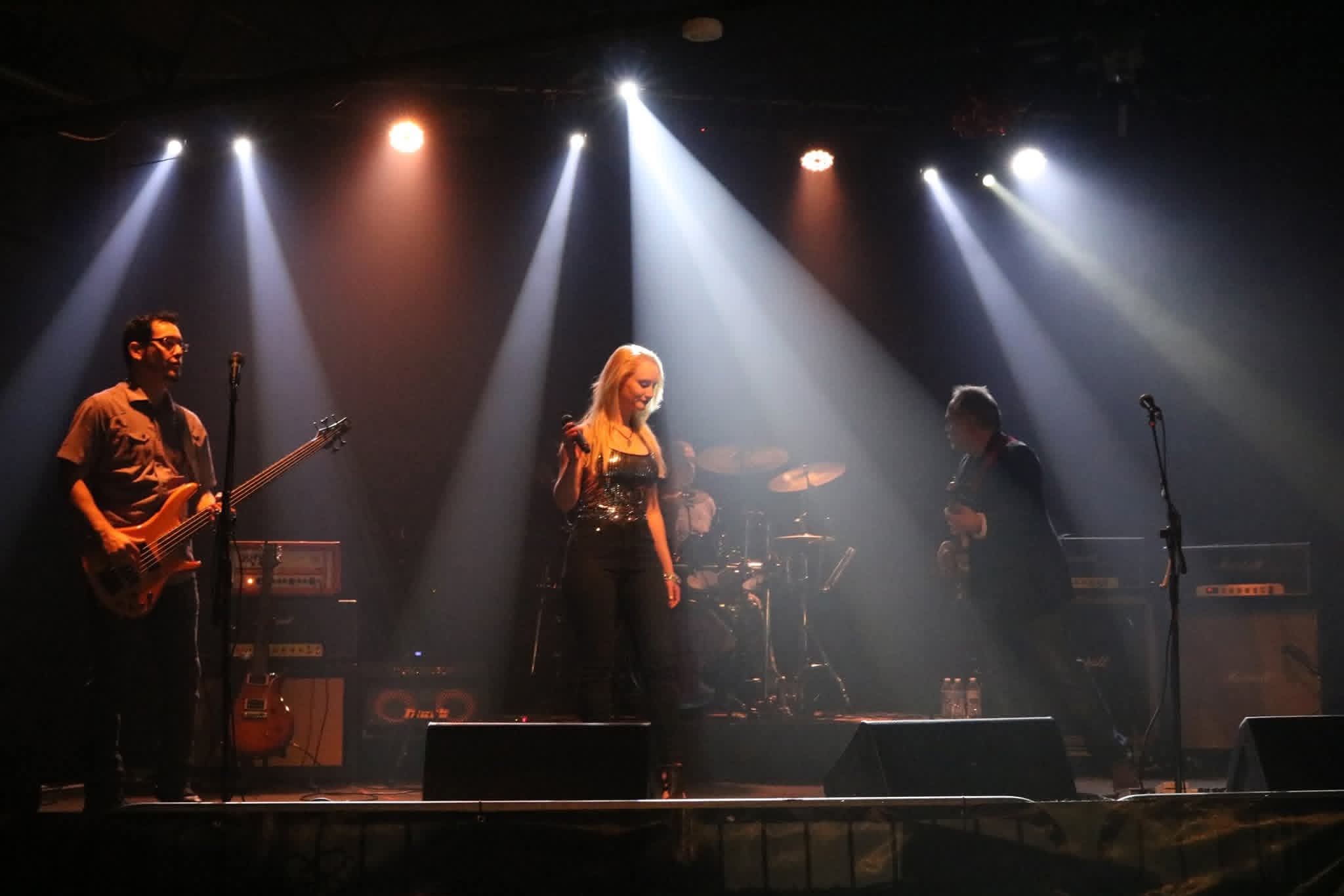 Three musicians perform on stage with bright spotlights overhead, playing instruments including guitar and drums.