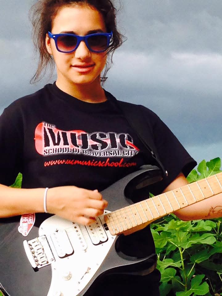 Young woman with sunglasses holding an electric guitar outdoors, cloudy sky in background.