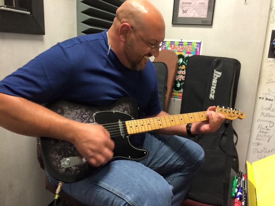 Man playing an electric guitar indoors, sitting on a chair, wearing glasses and a blue shirt.