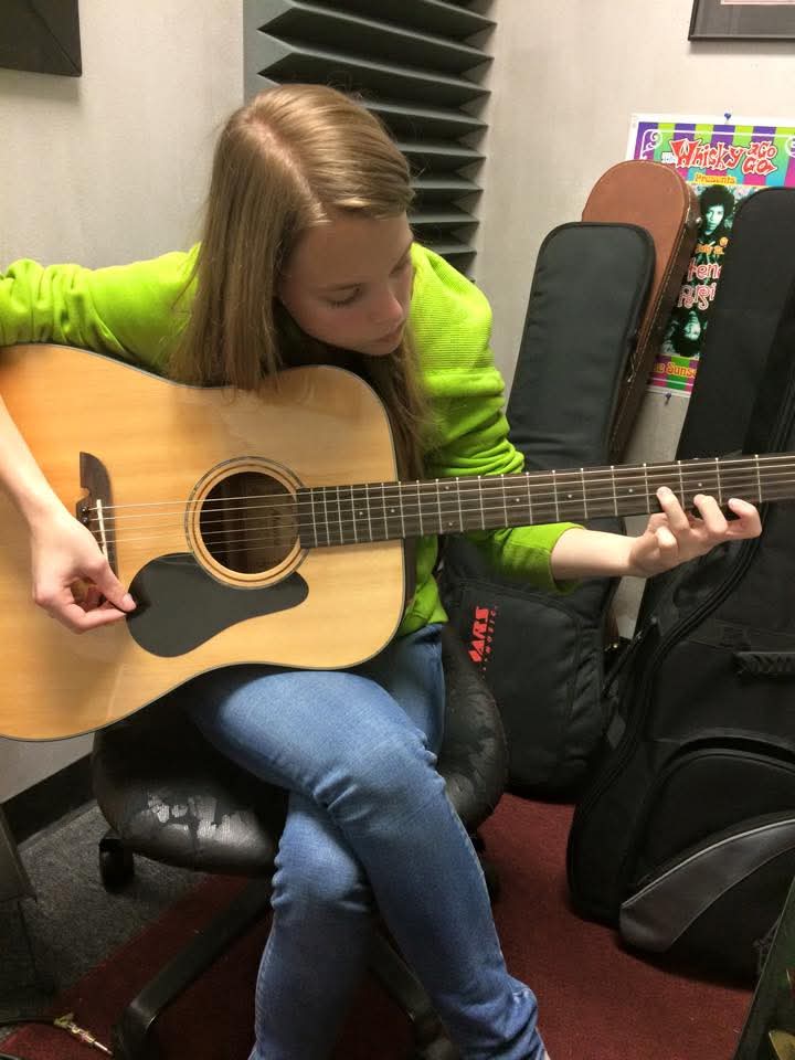 Young girl with long hair wearing a bright green sweater playing an acoustic guitar indoors.