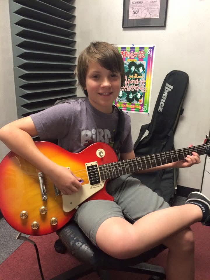 Child sitting on a chair holding an orange and yellow electric guitar, smiling, in a music room.