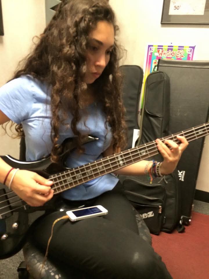 Young woman with long curly hair playing a bass guitar, sitting in a room with musical equipment and colorful posters.