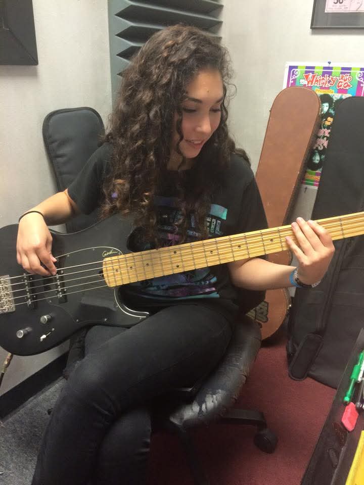 Young girl with curly hair playing an electric guitar in a room with chairs and colorful posters.