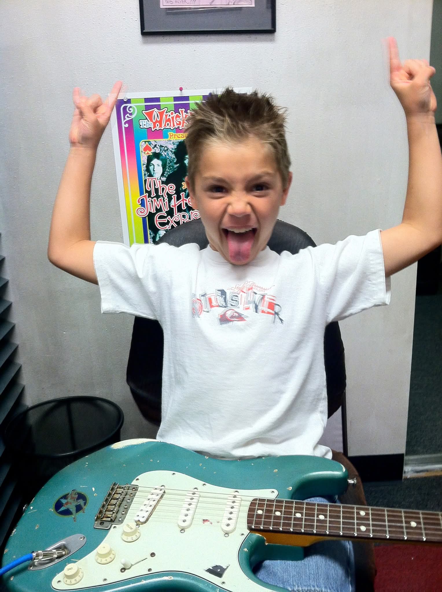 Child with arms raised, tongue out, smiling, sitting at a table with a guitar in front, celebrating.