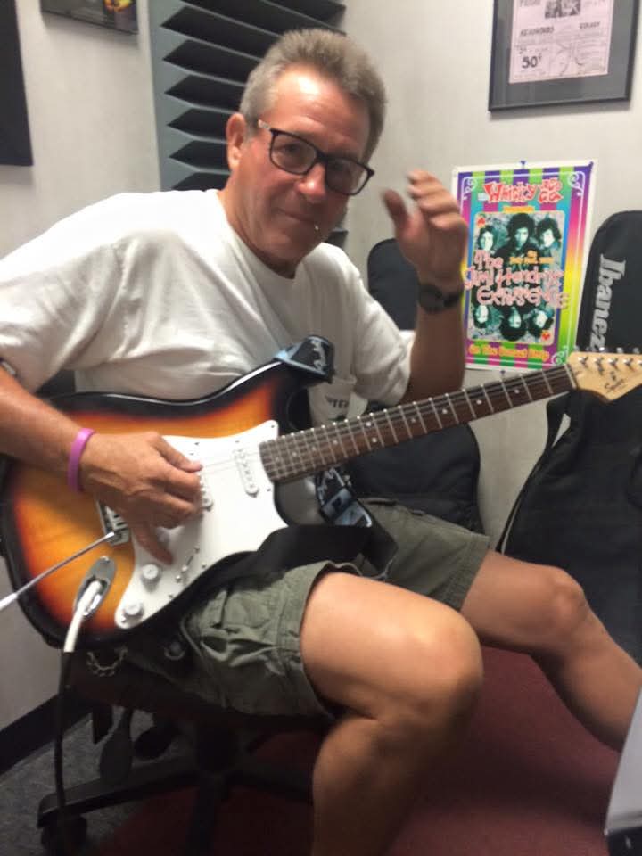 Person sitting in a chair holding an electric guitar, wearing glasses, a white shirt, and shorts, in a room with posters.