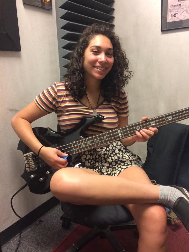 Young woman with curly hair smiling, sitting on a chair, holding an electric guitar, in a room with soundproofing panels.