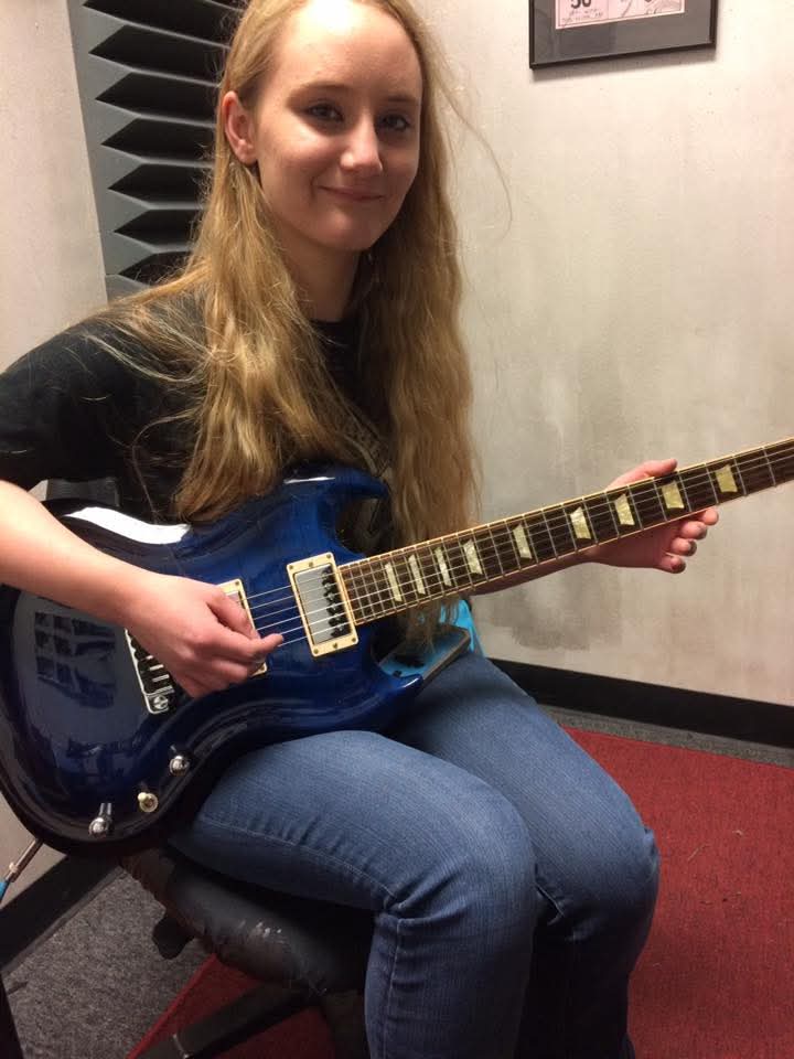 Young woman with long hair sitting on a chair, holding a blue electric guitar, smiling at the camera.